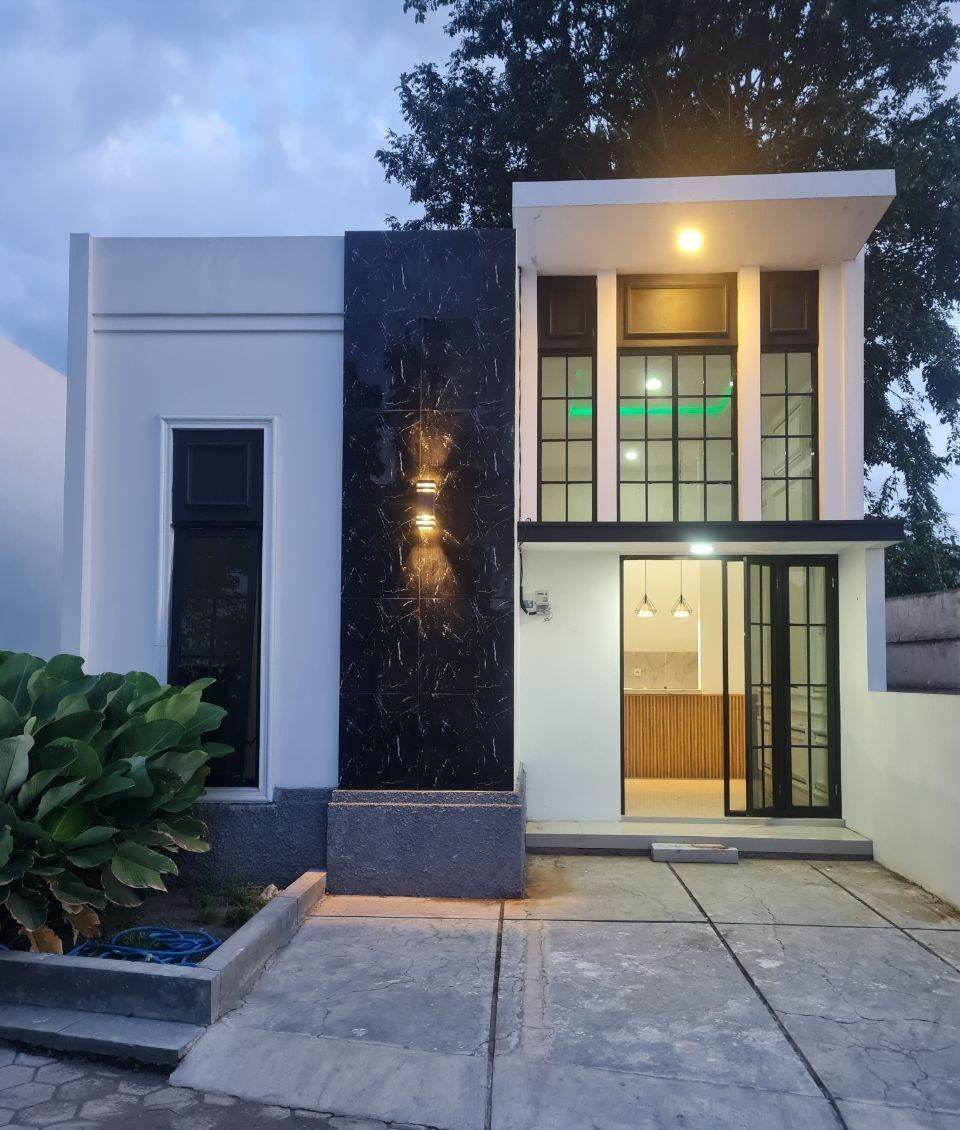 Rumah model Klasik Modern di Tirta Sepande Asri: fasad putih, panel granit hitam, jendela kaca tinggi berkusen hitam, foto sore hari dengan lampu teras menyala.